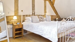 Un dormitorio con una estructura de cama de metal blanco, una mesita de noche de madera, una lámpara y un espejo. La habitación tiene vigas de madera a la vista en el techo.