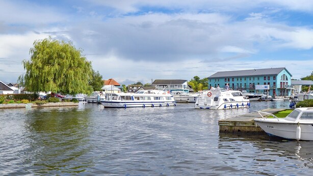 A calm river with boats docked along the shore, surrounded by residential buildings and a large tree.
