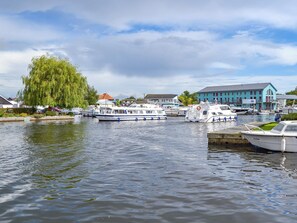 A calm river with boats docked along the shore, surrounded by residential buildings and a large tree. - Ideal for a couple or small family wishing to discover the Norfolk Broads. (Wroxham)