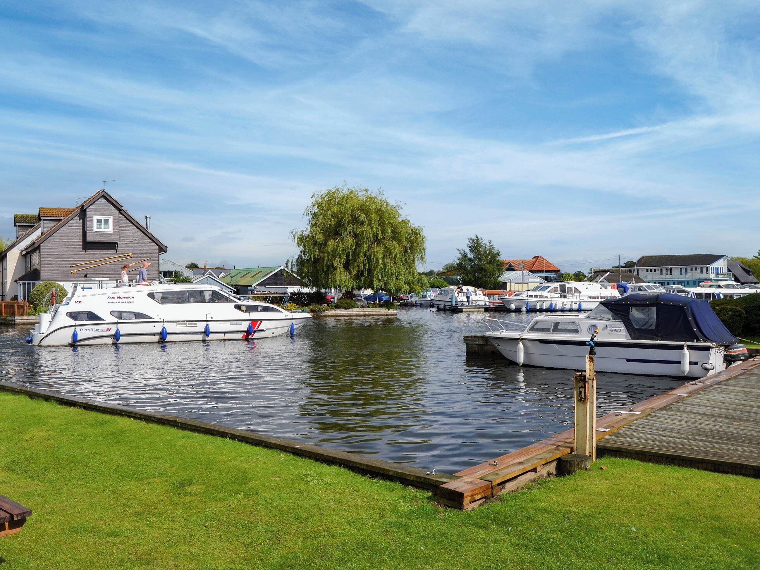 A marina with several boats docked, a grassy area in the foreground, and houses in the background.