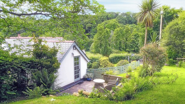 Una piccola casa bianca con tetto spiovente, circondata da vegetazione rigogliosa e un giardino curato.