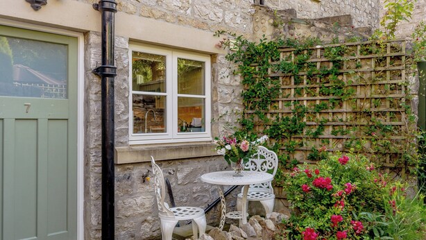A stone building with a green door, a window with a white frame, and a small white table with a chair. - 3 bedroom accommodation in Ashford-in-the-Water, near Bakewell (Ashford-in-the-Water, near Bakewell)