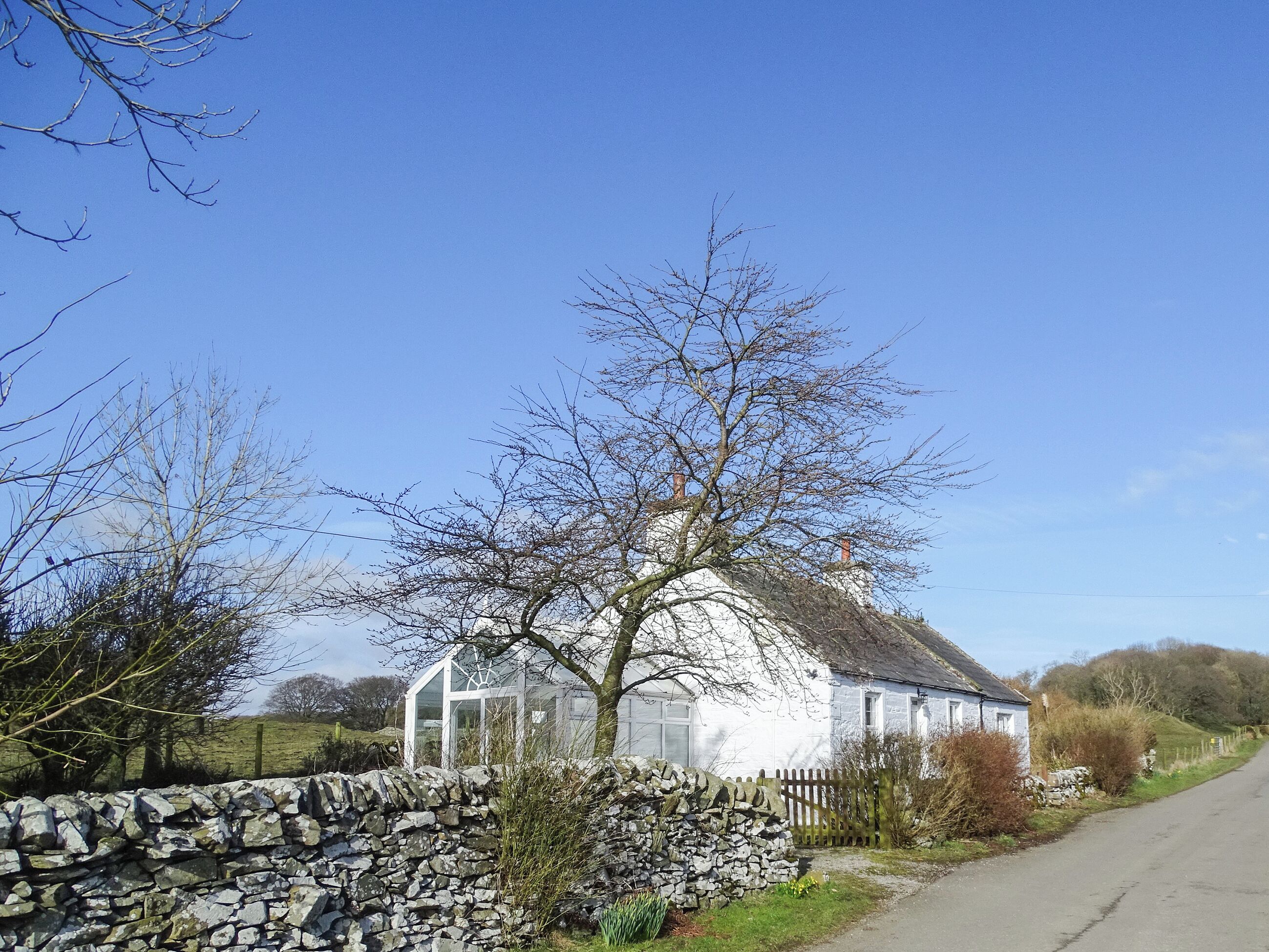 Un bâtiment blanc avec un mur en pierre et une clôture en bois, entouré d’arbres et sous un ciel dégagé.