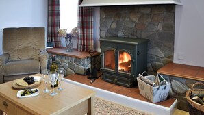 A cozy living room with a stone fireplace, a comfortable sofa, a wooden table with a cheese board and wine glasses, and a window with plaid curtains. - 2 bedroom accommodation in Urray, near Dingwall (Urray, near Dingwall)