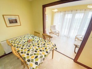 Un coin repas avec une table recouverte d’une nappe à motifs de feuilles, des chaises en bois et une vue sur un salon où l’on aperçoit un fauteuil blanc et une fenêtre avec des rideaux.