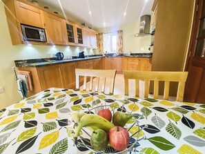 A kitchen with wooden cabinets, a microwave, a dining table with a fruit bowl, and a window with curtains. - 3 bedroom accommodation in Bamburgh (Bamburgh)