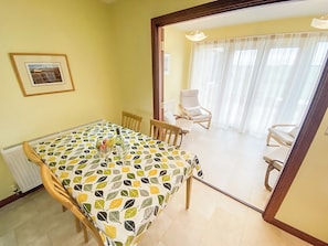 A dining area with a table covered in a leaf-patterned tablecloth, wooden chairs, and a view into a living area with a white chair and a window with curtains.