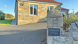 A stone cottage with a tiled roof, a paved driveway, and a sign indicating the name 'Friarystone Cottage.'