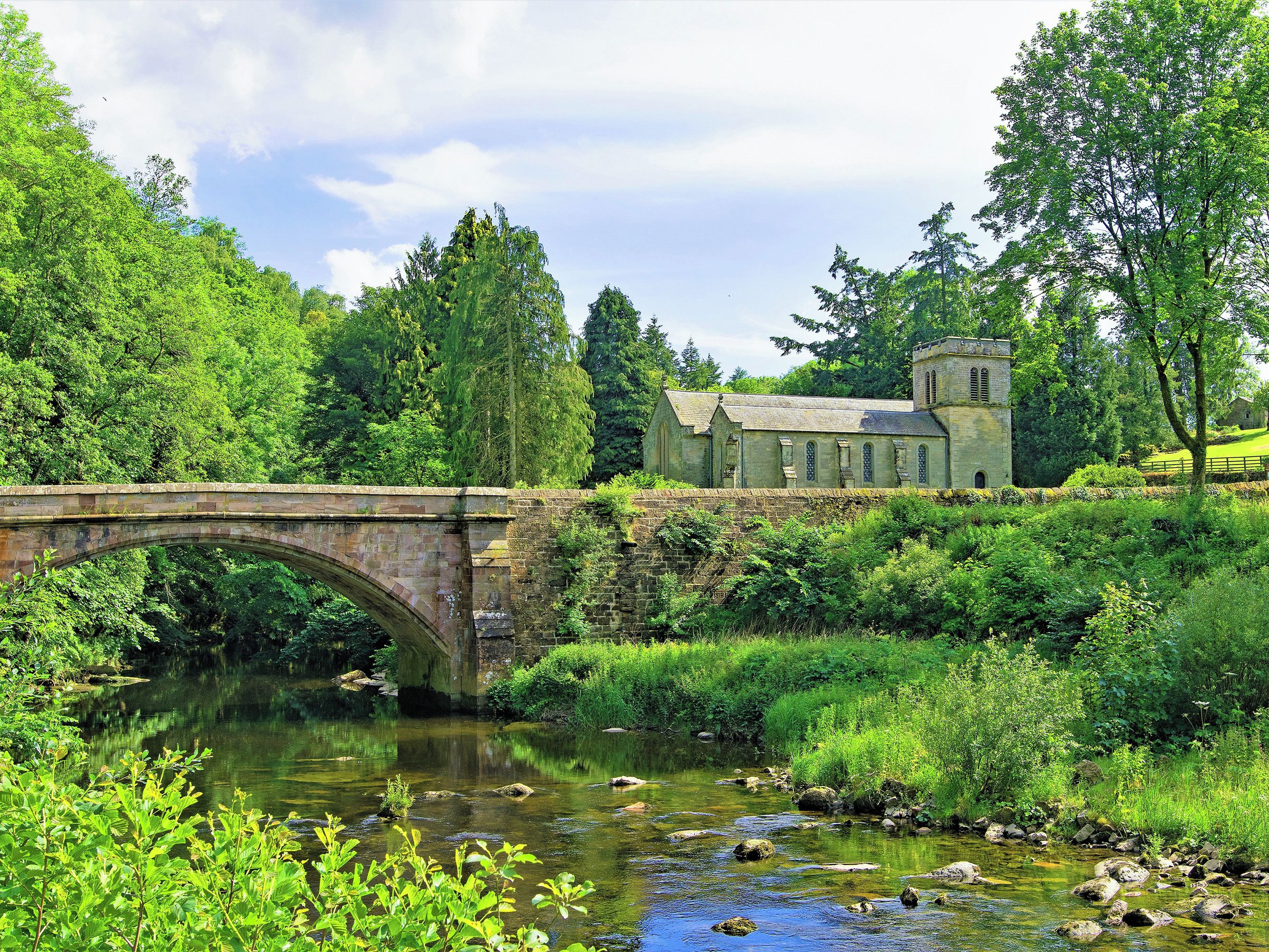 A stone bridge over a calm river, surrounded by lush greenery and a historic stone building with a tower.