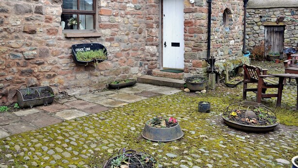 A stone building with a white door, a small window, and a stone wall. There are potted plants and a fire pit in the courtyard. - 3 bedroom accommodation in Crackenthorpe, Appleby (Crackenthorpe, Appleby)