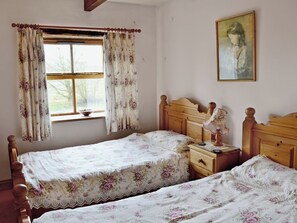 A bedroom with two single beds, a wooden headboard, a bedside table with a lamp, and a window with floral curtains.