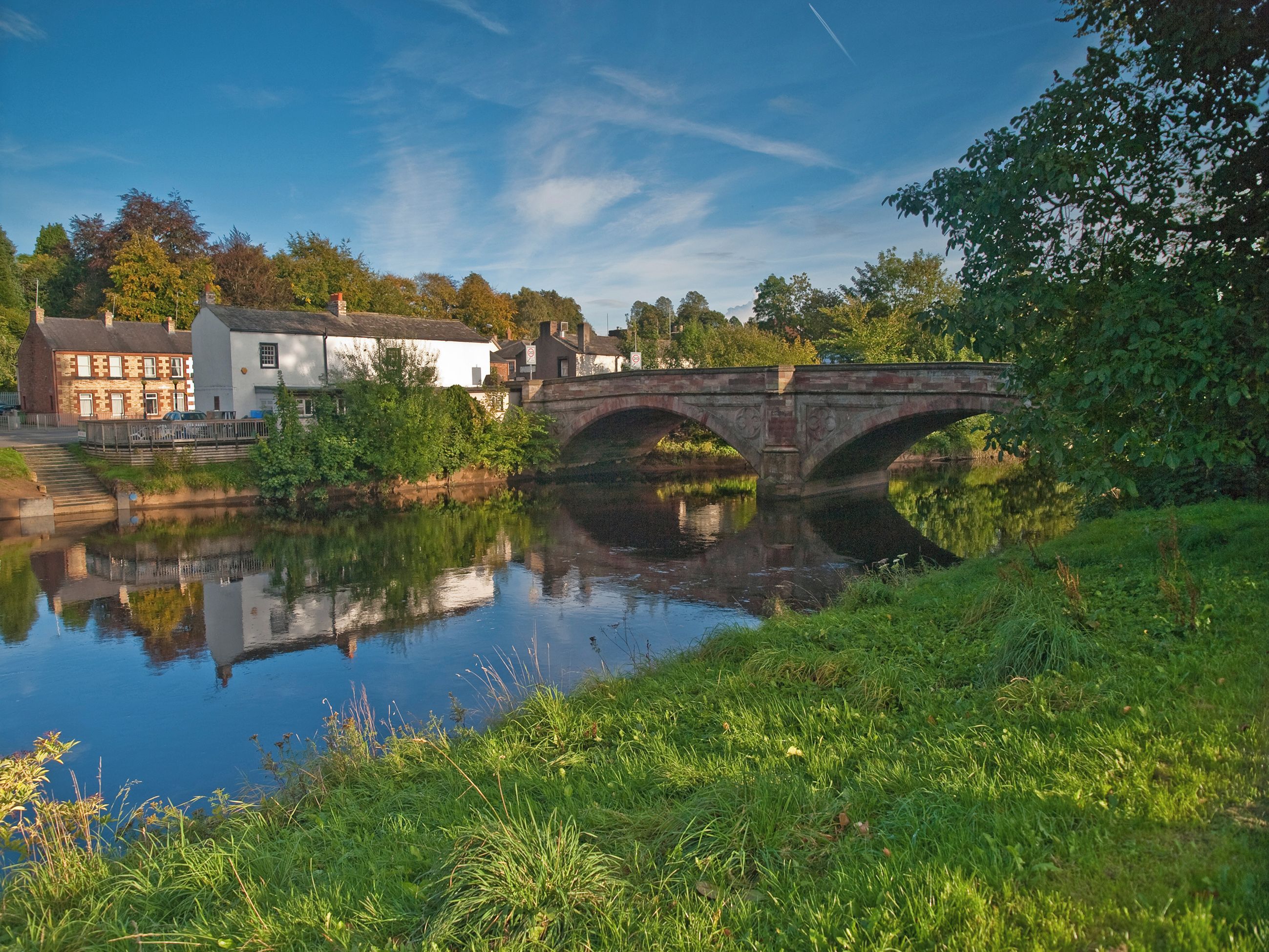 A stone bridge over a calm river, surrounded by greenery and buildings.