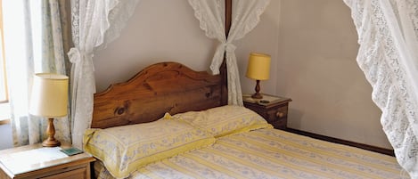 Quarto com cabeceira de madeira, cama com dossel, duas mesas de cabeceira com luminárias e um criado-mudo com abajur.