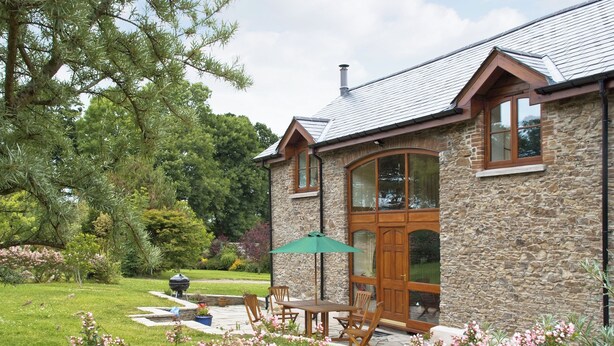 Une maison en pierre avec une grande fenêtre, une porte et une terrasse avec des chaises et une table.