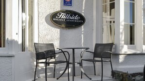 A sign for Hillside Holiday Apartments with a self-catering symbol, two chairs, and a small table outside. - 2 bedroom accommodation in Keswick (Keswick)