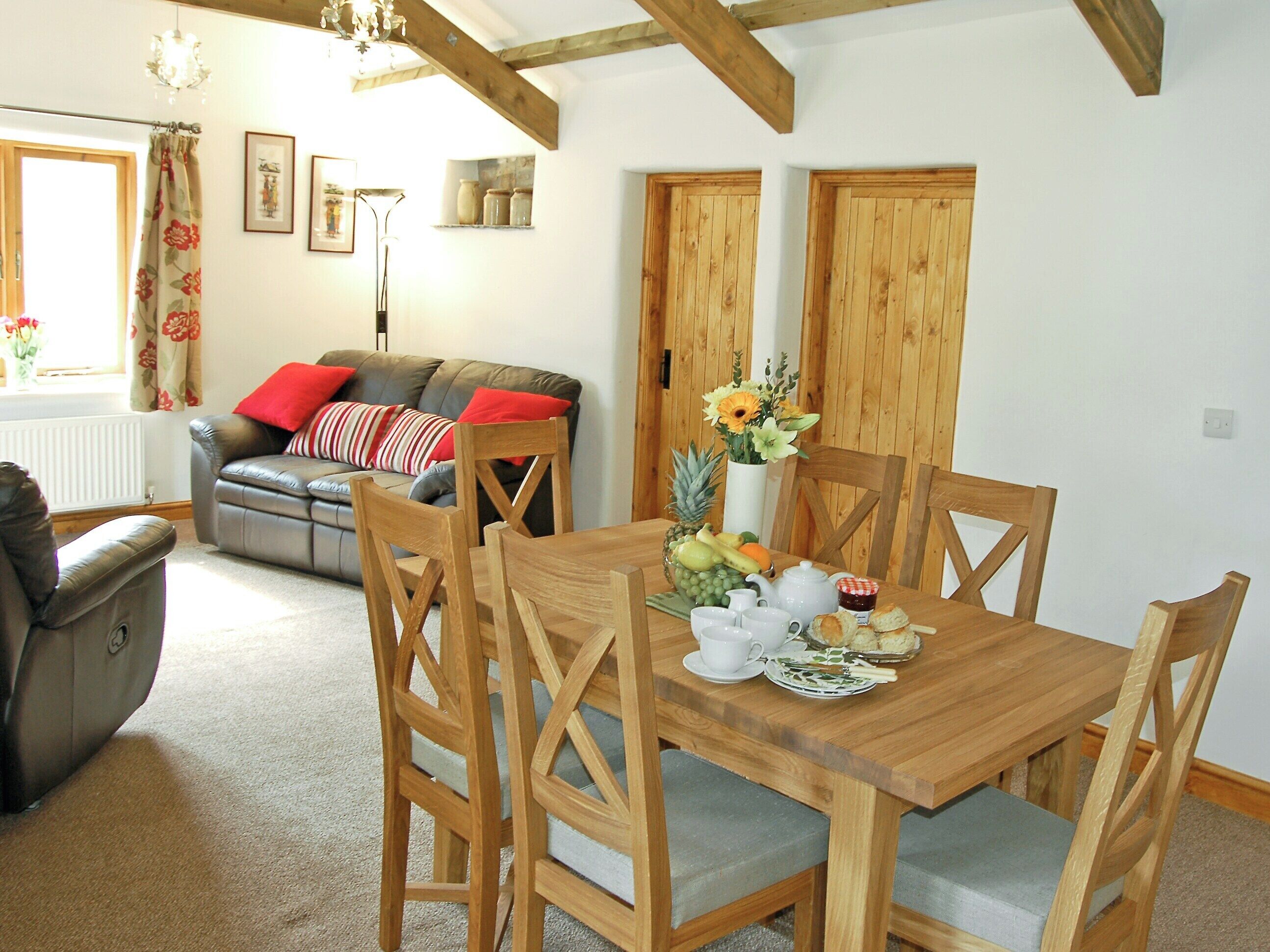 Un coin repas avec une table en bois dressée pour quatre personnes, entourée de chaises en bois dotées d’assises rembourrées. Un arrangement floral orne la table, et un panier de fruits est visible.