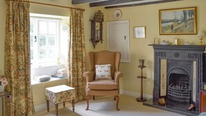 A room with a traditional fireplace, a wooden chair with a cushion, a small table, and a window with curtains.