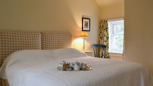 A large bed with a tray of tea and biscuits, a checkered sofa, a lamp, and a chair by a window.