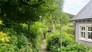 Camí de jardí que condueix a una casa, amb una font visible i vegetació abundant.