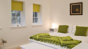 Un dormitorio con dos ventanas, una mesita de noche con lámpara, cama con ropa de cama verde y un cuadro enmarcado en la pared.