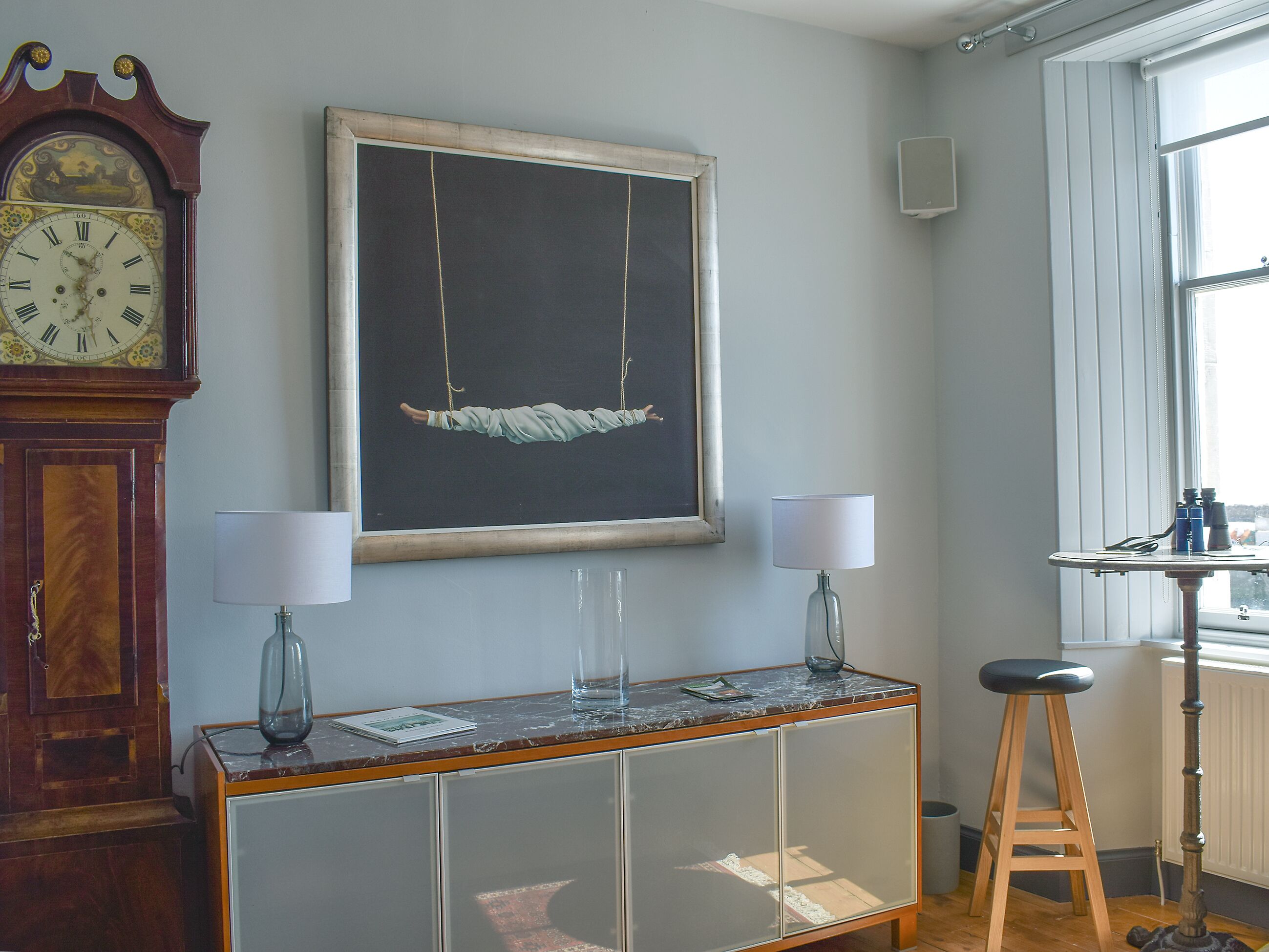 A room with a wooden cabinet, two lamps, a framed artwork, a large window, and a wall-mounted clock.