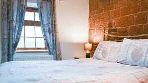 Un dormitorio con pared de ladrillo, una ventana con cortinas, una cama con ropa de cama estampada y una lámpara de noche.