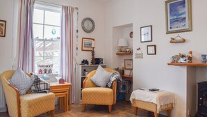 A cozy living room with two armchairs, a small table, and a sofa. There is a window with curtains, a clock on the wall, and various decorations including framed pictures and a small shelf with items.