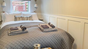 Una cama bien hecha con un edredón gris acolchado, dos toallas dobladas, una bandeja con una cafetera y tazas, y una ventana con vista a una valla rosa.