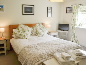 A neatly made bed with floral pillows, a bedside lamp, a television on a small table, and a window with curtains.
