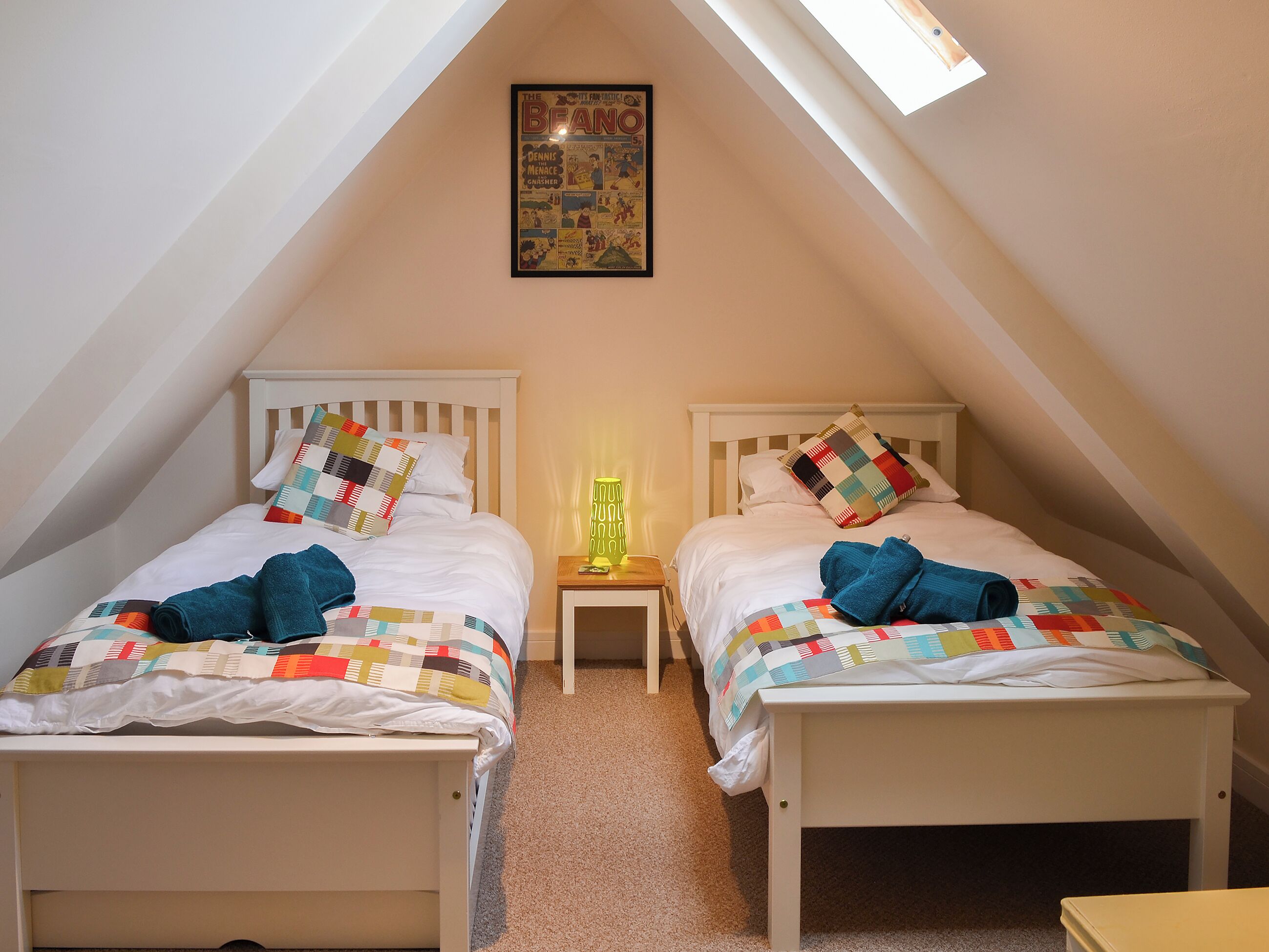 A room with two beds, a small bedside table with a lamp, a framed comic strip on the wall, and a sloped ceiling.