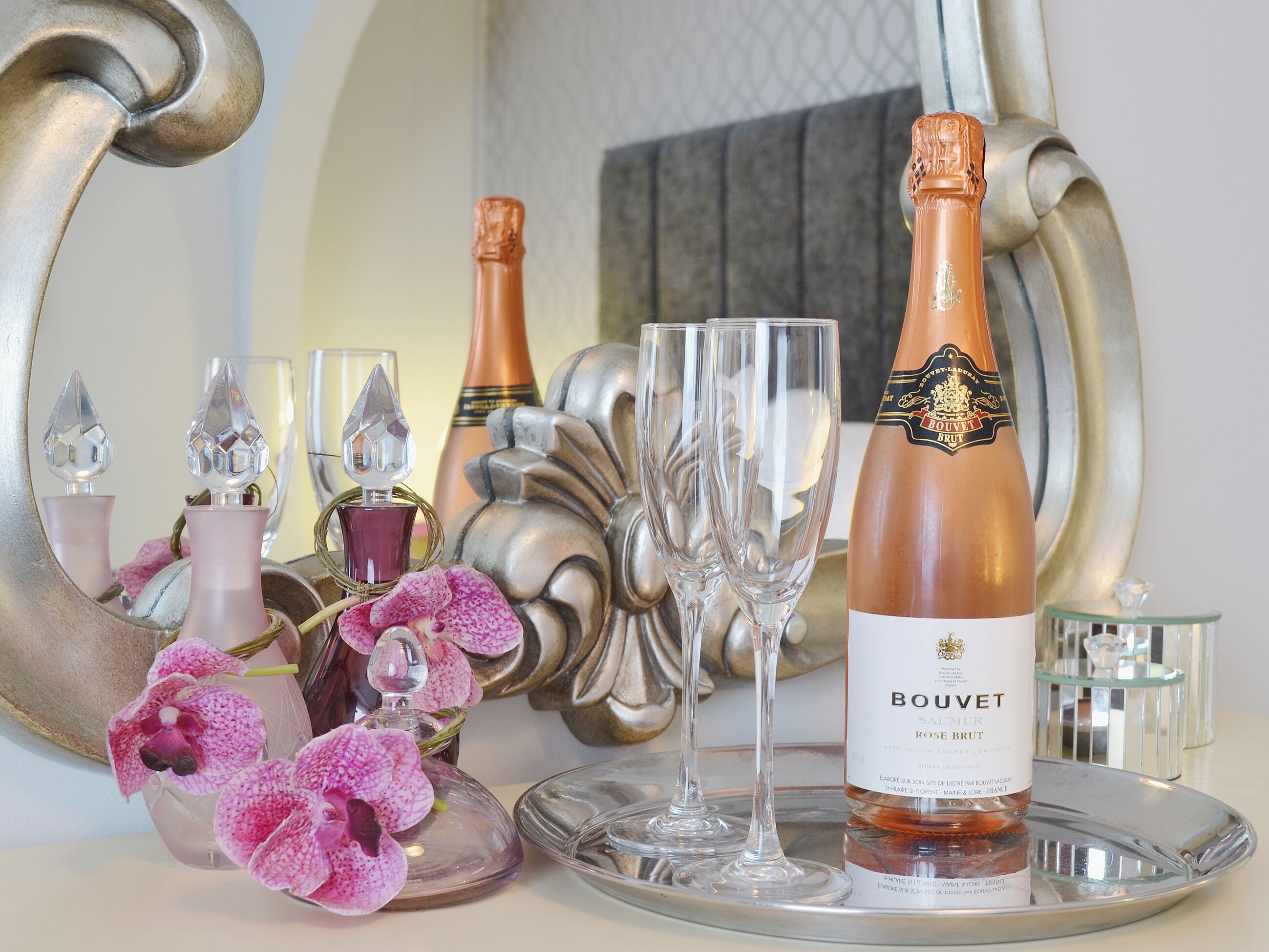 Une bouteille de champagne BOUVET SAUTER ROSE BRUT, deux verres à vin à pied transparents, un plateau décoratif en argent et des fleurs d’orchidée roses.