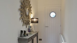 A hallway with a white door, a round window, a wall-mounted mirror, a table with decorative items, and a floor lamp. - 2 bedroom accommodation in Aberystwyth (Aberystwyth)