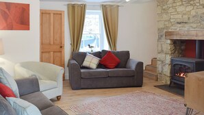 A cozy living room with a stone fireplace, a grey sofa, a wooden door, and a window with curtains. - 3 bedroom accommodation in Llanarth, near New Quay (Llanarth, near New Quay)