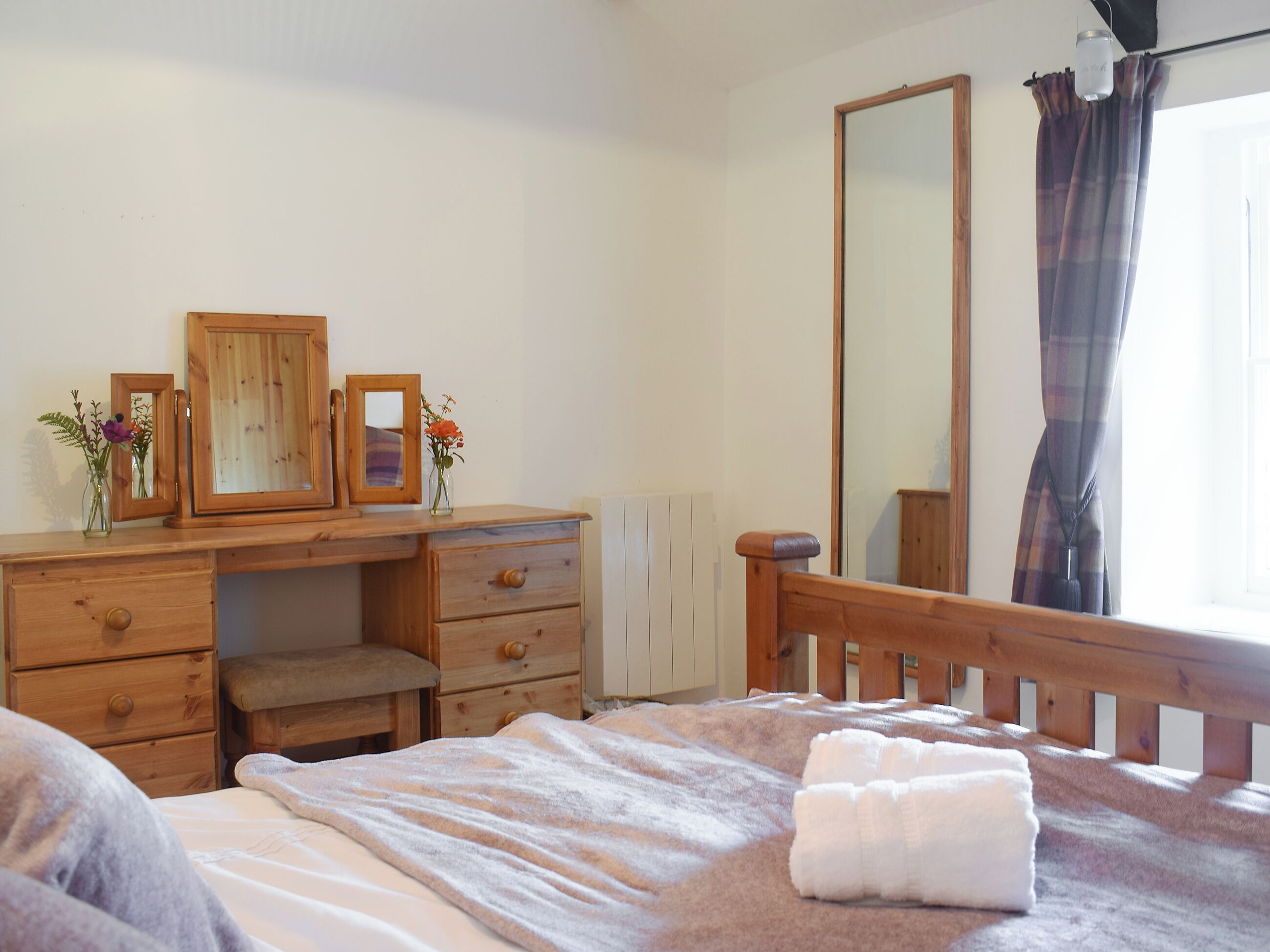 Een slaapkamer met een houten dressoir, een spiegel, een bed met een witte deken en een raam met gordijnen.