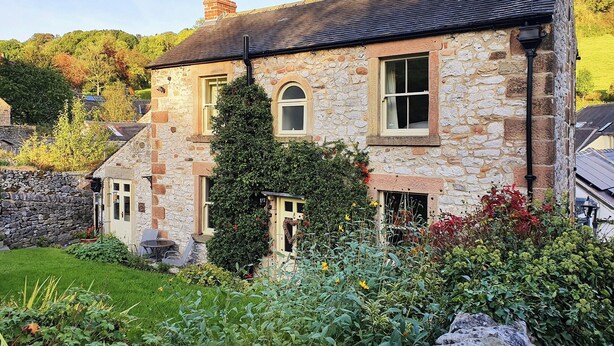 Une maison en pierre avec un toit en pente, une cheminée et un jardin où poussent différentes plantes et fleurs.