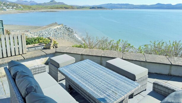 A patio with wicker furniture, a table, and an umbrella, overlooking a body of water and distant mountains. - 3 bedroom accommodation in Criccieth, near Porthmadog (Criccieth, near Porthmadog)