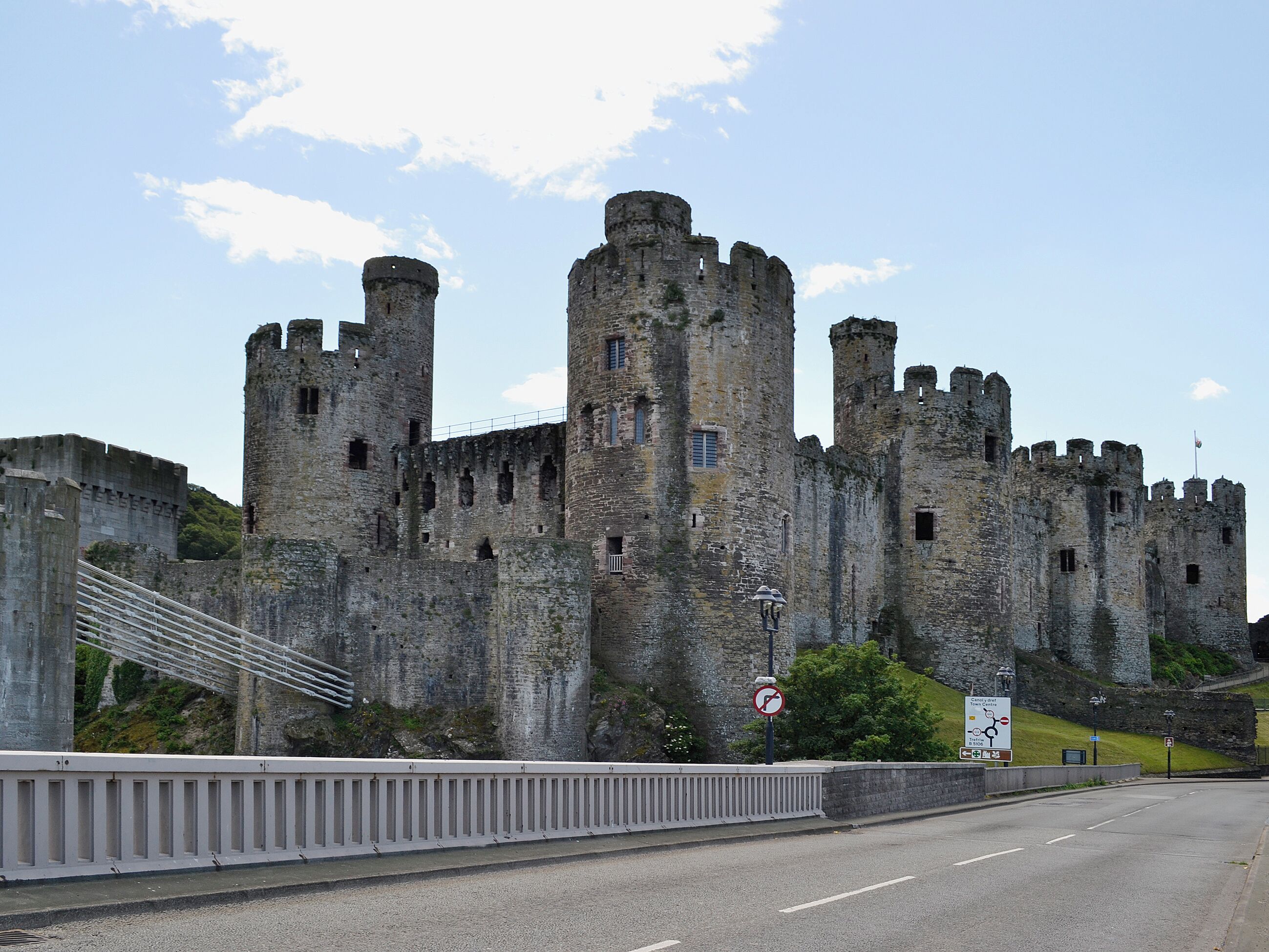 A historic castle with multiple towers, a bridge in the foreground, and a sign indicating a parking area.