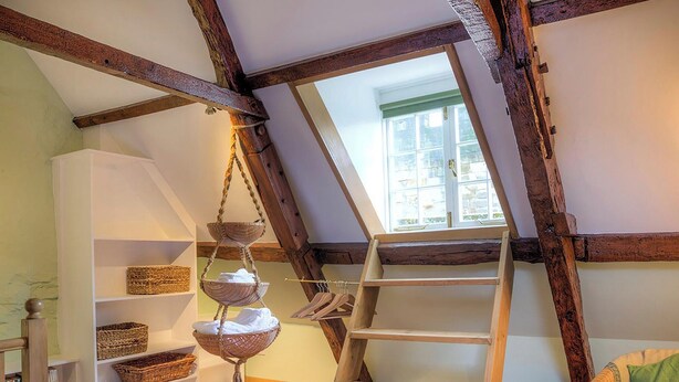 Quarto no estilo loft com vigas de madeira aparentes, teto inclinado e claraboia. Há prateleiras com cestos e um cesto suspenso com toalhas.