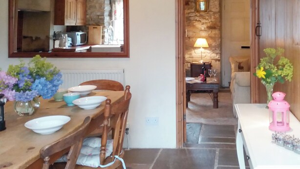 Un coin repas avec une table en bois, des chaises et un miroir au-dessus. On peut apercevoir une cuisine à travers une porte ouverte.