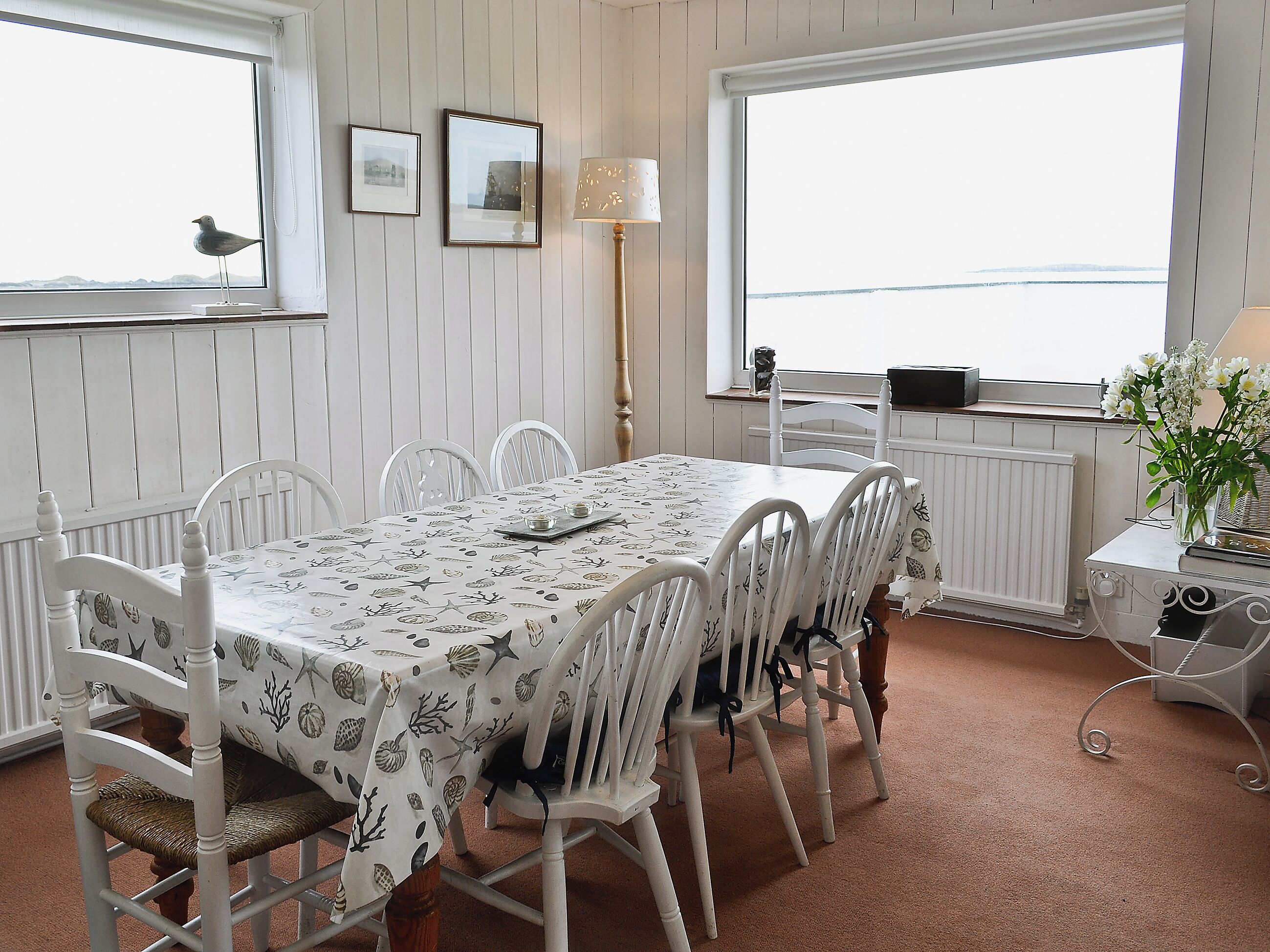 Un coin repas avec une table et des chaises blanches, des tableaux encadrés au mur et une vue sur la mer par de grandes fenêtres.