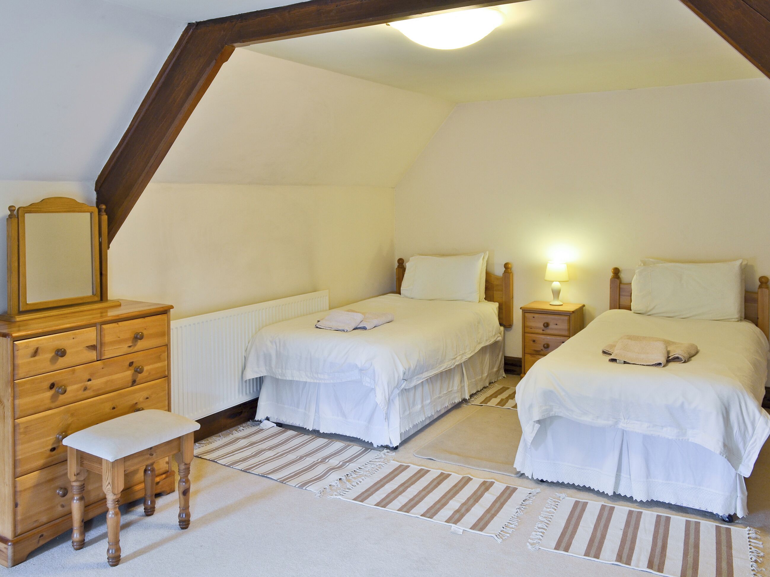 A bedroom with two beds, a wooden dresser, a bench, and a nightstand with a lamp.