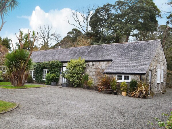 A stone house with a gravel driveway, surrounded by trees and plants.