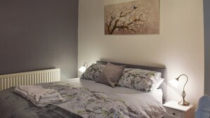 Un lit bien fait, avec une literie à motifs floraux, une lampe de chevet et un tableau représentant des oiseaux perchés sur un arbre.