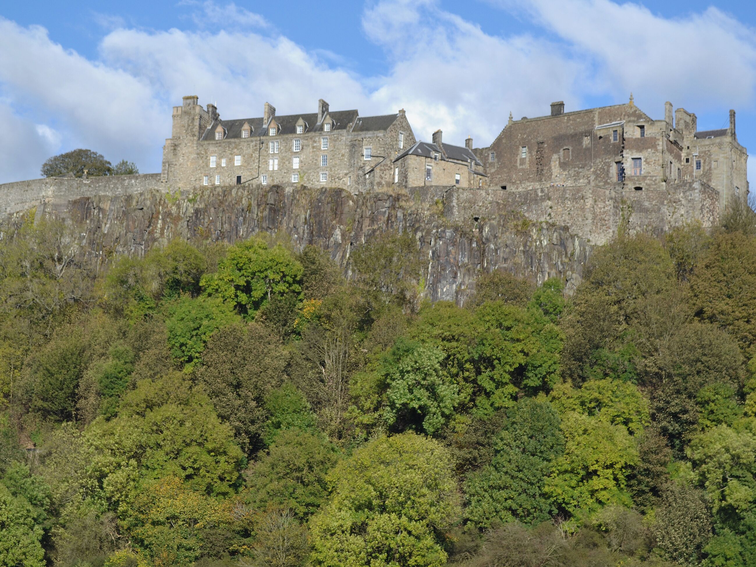 A historic stone castle with multiple buildings atop a cliff, surrounded by dense green foliage.