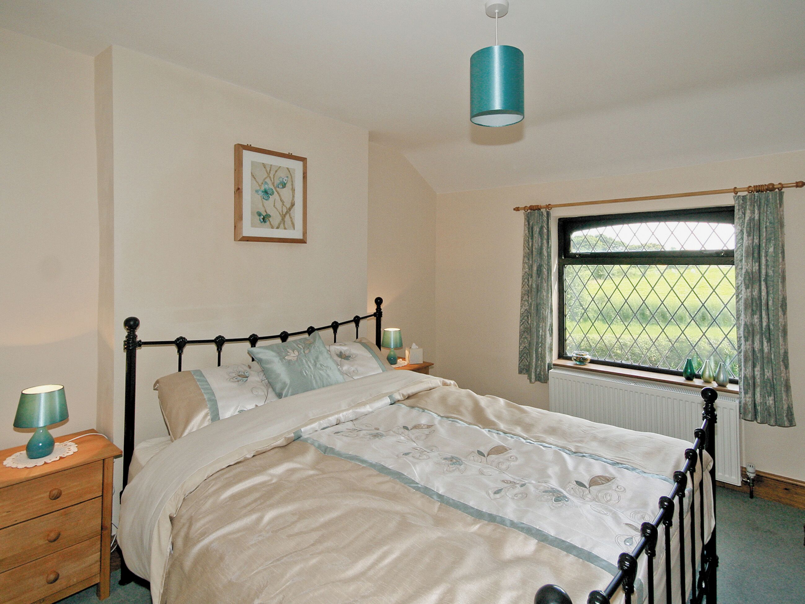 A bedroom with a bed, bedside tables, a window with curtains, a radiator, and a hanging light fixture.