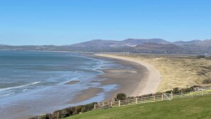 A coastal landscape with a sandy beach, grassy areas, and distant mountains.