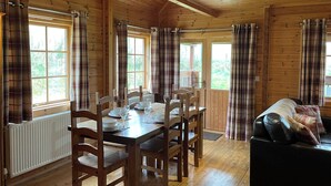 A wooden cabin interior with a dining table, chairs, a black leather sofa, plaid curtains, and a window with a view of greenery.