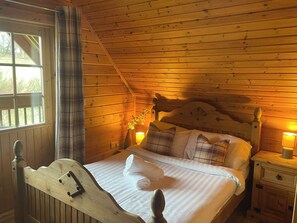 A wooden cabin bedroom with a bed, pillows, a nightstand, and a window. - Sun Rise Lodge No 10 - HW7655 (Llanbedr)