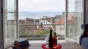 A room with a large window overlooking a coastal view, a table with books and wine bottles, and a chair. - 4 bedroom accommodation in St Monans, near Anstruther (St Monans, near Anstruther)