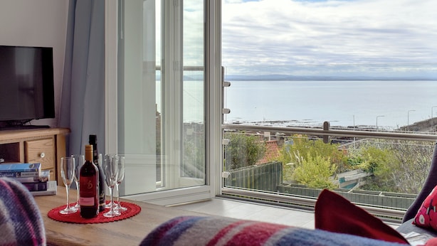 A room with a TV, a table with wine glasses and a bottle, and a large sliding glass door leading to a balcony with a view of the sea. - 4 bedroom accommodation in St Monans, near Anstruther (St Monans, near Anstruther)